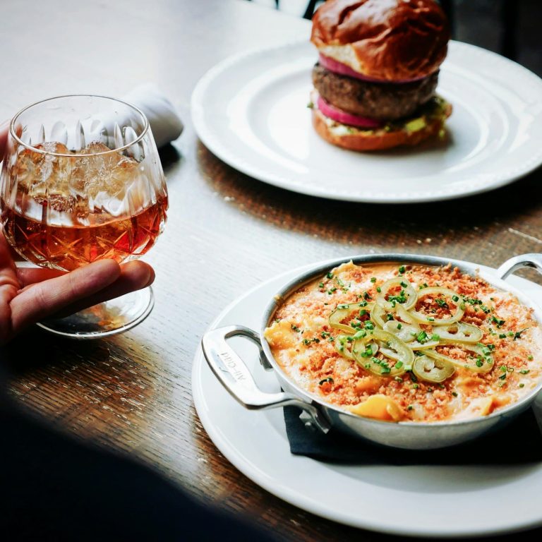 Hand met glas whiskey naast een gerecht en een hamburger als onderdeel van bewust eten in december.